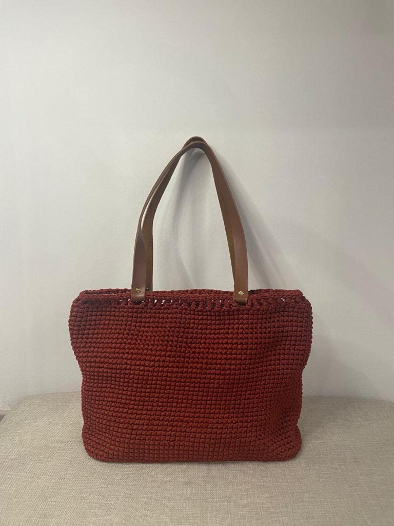 Grand sac à main cabas en crochet de couleur rouge brique terracotta avec finition maille serrée. Le sac dispose de deux longues anses camel pour un porté épaule. Création artisanale française Vanda, posée sur un support neutre.