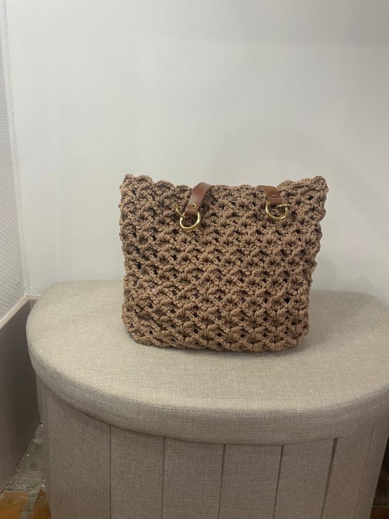 Sac à main type cabas en crochet de couleur marron moka avec un point texturé en relief. Le sac possède des anses camel attachées par des anneaux dorés. Création artisanale française posée sur un pouf beige dans la boutique de Vanda à Albi.
