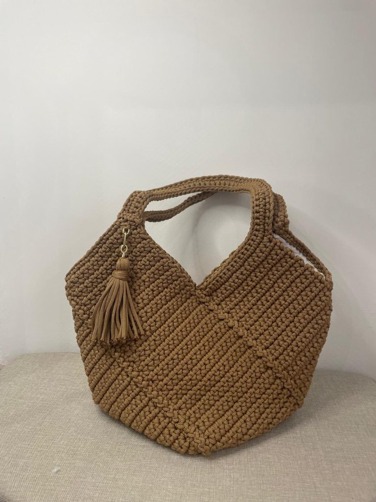 Sac à main en crochet de couleur caramel avec une forme géométrique en V style origami. Les anses sont entièrement crochetées dans la même matière. Un pompon décoratif est attaché sur le côté. Création artisanale Vanda posée sur un support neutre.