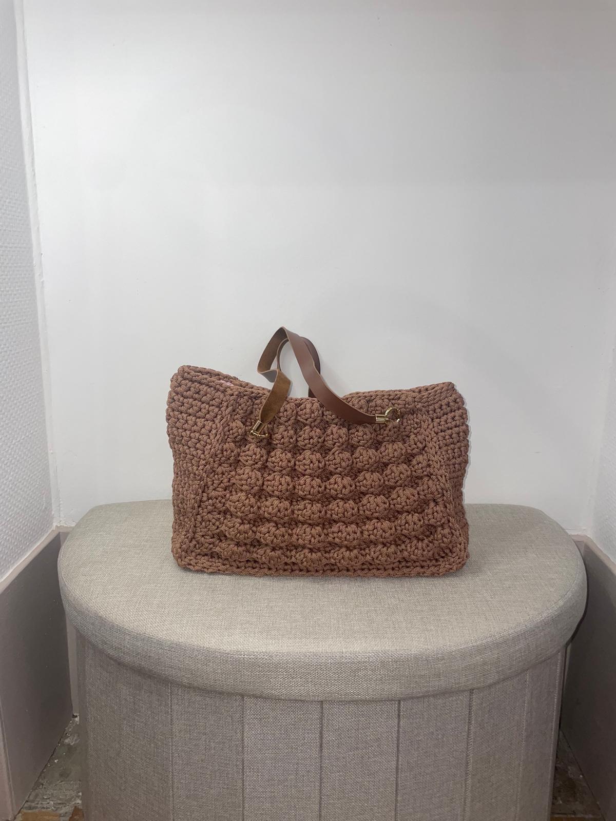 Sac à main en crochet couleur marron praliné avec un point texturé style noisette ou popcorn en relief sur le devant. Anses marron avec anneaux dorés. Création artisanale Vanda posée sur un pouf dans la boutique d'Albi.