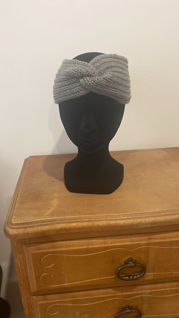 Bandeau d'hiver pour femme en crochet gris souris avec torsade sur le devant (style turban). Accessoire cheveux chaud et mode présenté sur une tête de mannequin noir. Création artisanale fait main par Vanda Albi