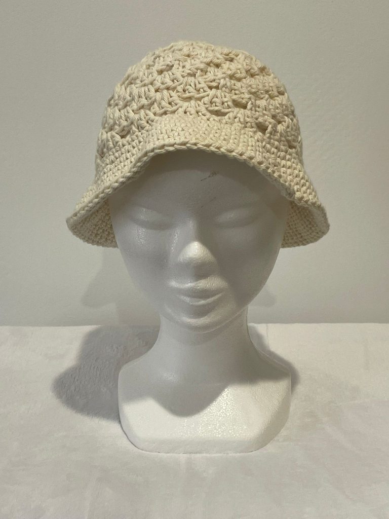 Chapeau type Bob pour femme en crochet de couleur écru naturel. Le haut du chapeau présente un point fantaisie texturé et aéré, tandis que le bord est en mailles serrées. Présenté sur une tête de mannequin blanc. Création artisanale d'été Vanda.