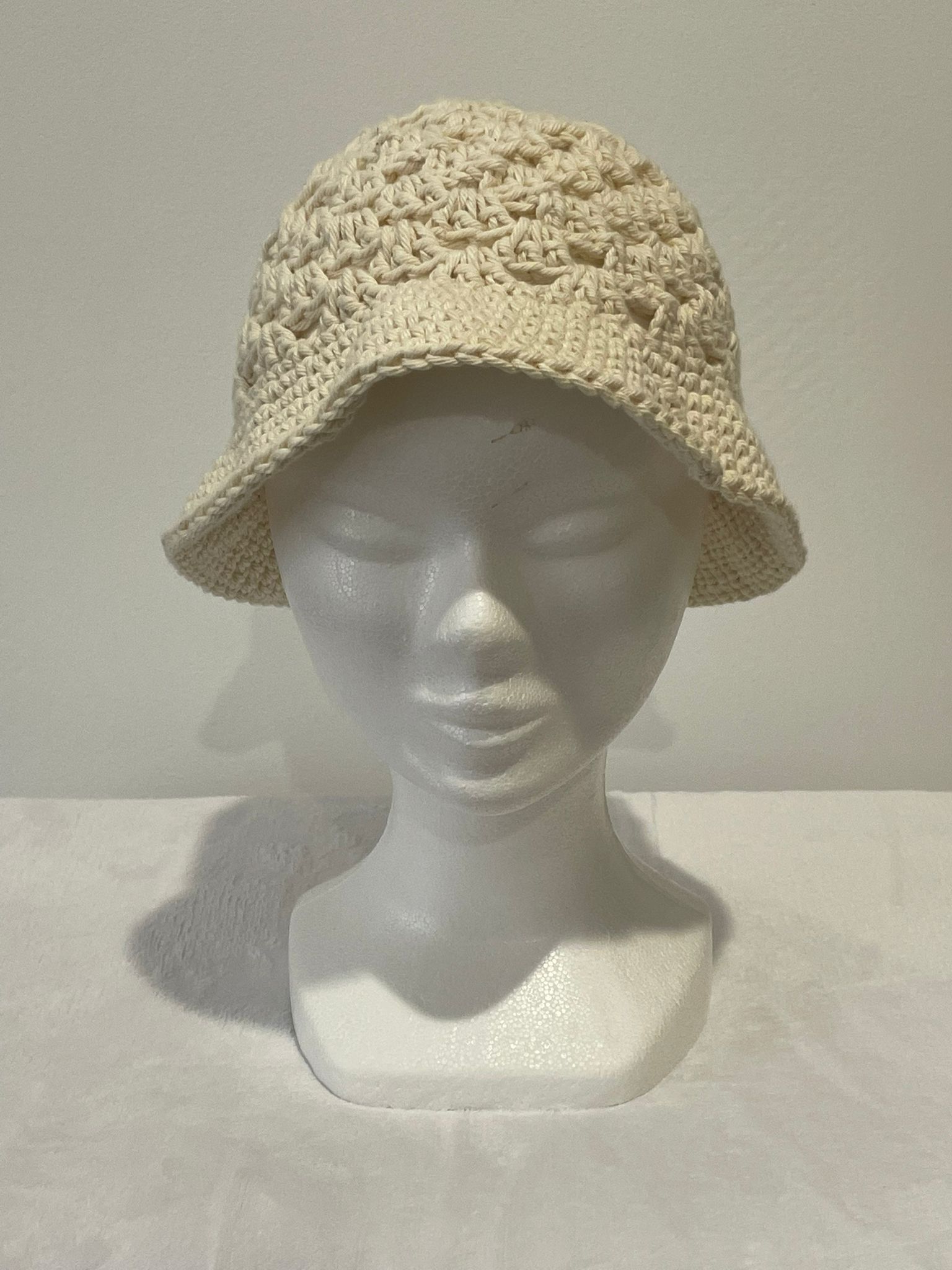 Chapeau type Bob pour femme en crochet de couleur écru naturel. Le haut du chapeau présente un point fantaisie texturé et aéré, tandis que le bord est en mailles serrées. Présenté sur une tête de mannequin blanc. Création artisanale d'été Vanda.