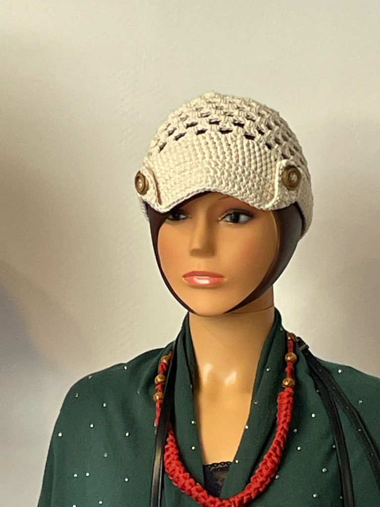 Casquette femme en crochet de couleur écru beige avec une visière rigide et un corps en mailles ajourées. Deux boutons décoratifs sont placés sur les côtés au-dessus de la visière. Présentée sur un mannequin portant également un collier rouge en crochet. Création artisanale française Vanda.