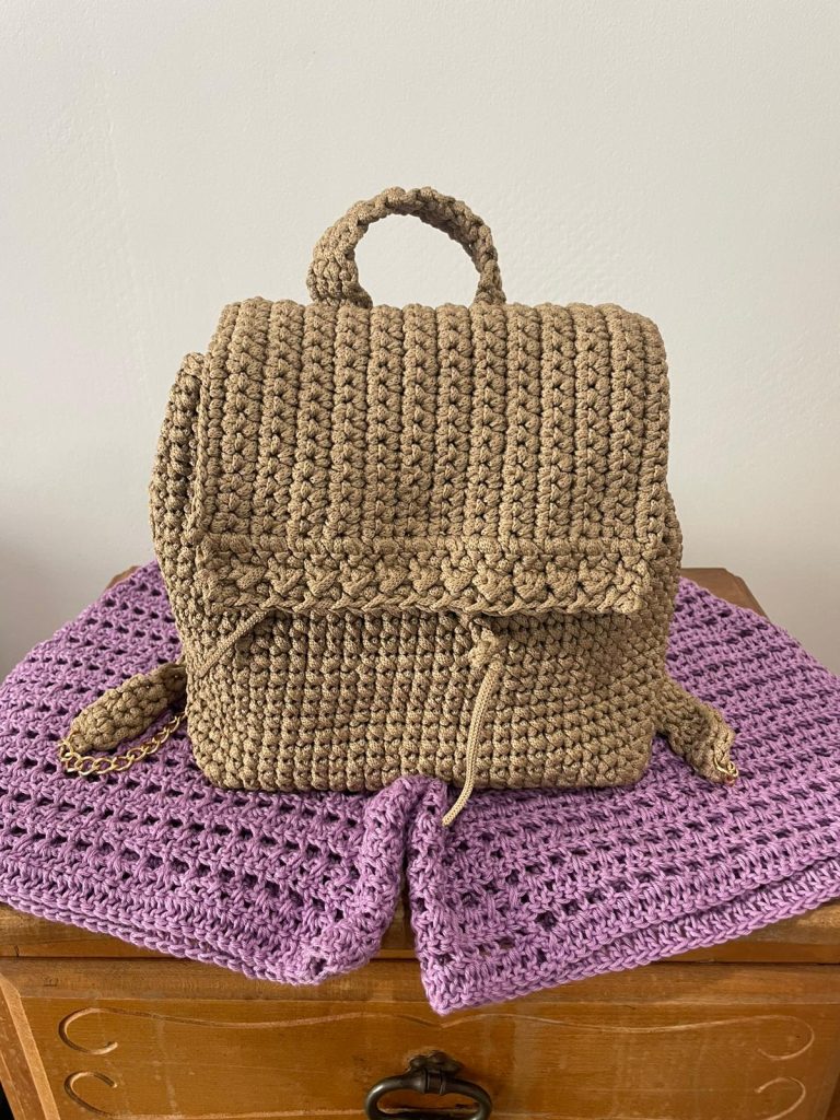 Petit sac à dos pour femme en crochet fait main de couleur taupe beige. Le sac possède un rabat texturé, une anse supérieure en crochet et des bretelles chaînette dorée. Posé sur un napperon violet. Création maroquinerie textile artisanale Vanda.