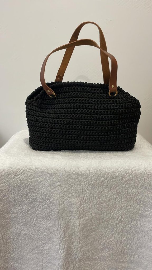 Sac à main en crochet de couleur noire intense avec une maille serrée et structurée. Le sac dispose de deux anses courtes couleur camel attachées par des anneaux métalliques dorés. Création artisanale française Vanda posée sur fond blanc.
