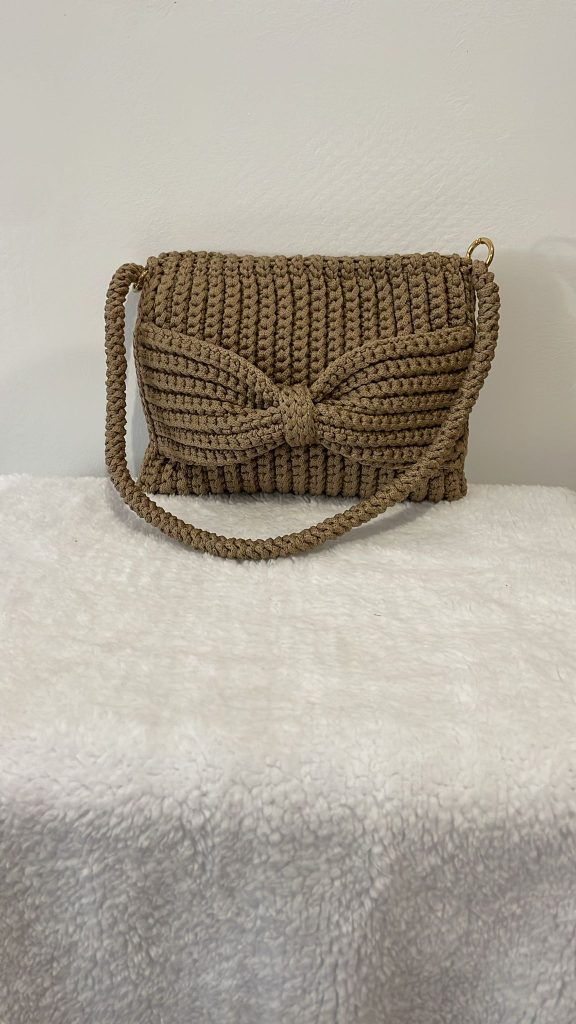 Sac à main rectangulaire en crochet de couleur beige sablé avec un grand nœud papillon en relief sur le devant. Le sac possède une texture nervurée verticale et une bandoulière en cordelette de crochet assortie. Création artisanale féminine Vanda.