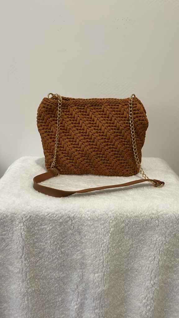 Sac à main en crochet couleur caramel avec un motif texturé en forme d'épi ou de chevron. Le sac dispose d'une bandoulière en chaîne dorée avec un renfort pour l'épaule assorti. Création artisanale élégante Vanda sur fond blanc.