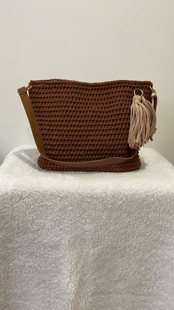 Sac à main en crochet couleur marron chocolat posé sur un fond blanc. Le sac est orné d'un gros pompon (tassel) décoratif beige et taupe sur le côté et possède une bandoulière marron assortie. Création artisanale française Vanda à Albi.