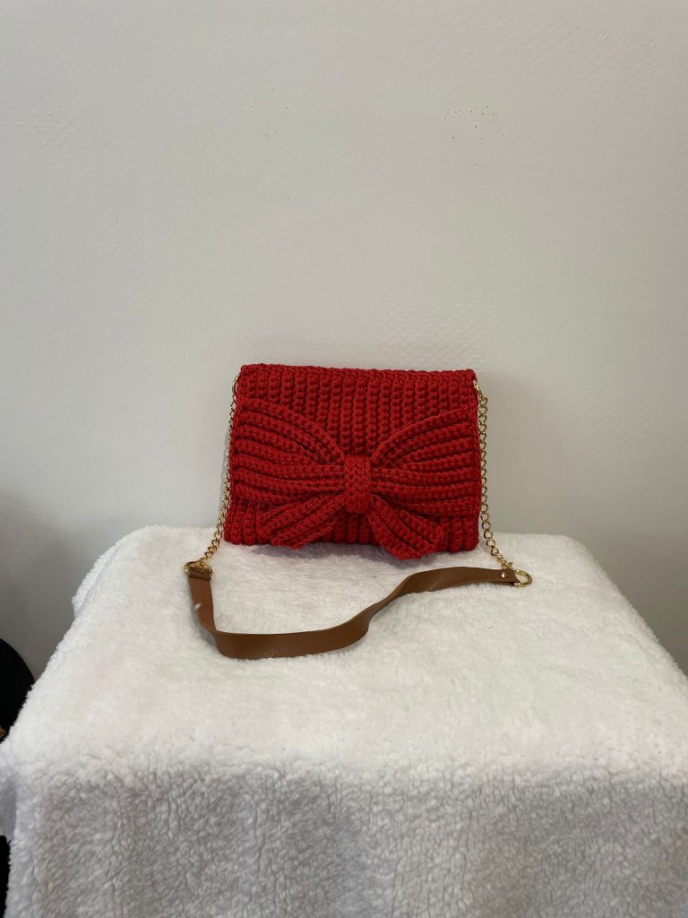 Sac à main rectangulaire en crochet de couleur rouge vif, orné d'un grand nœud papillon en relief sur le devant. Le sac possède une bandoulière en chaîne dorée avec un renfort épaule contrastant. Création artisanale glamour Vanda sur fond blanc.