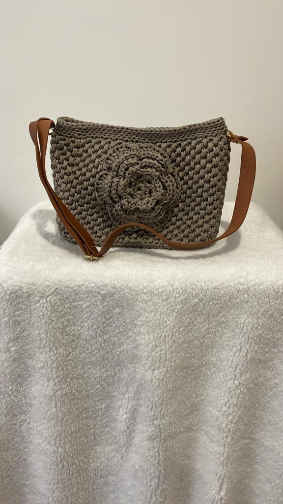 Sac à main en crochet de couleur taupe/grège orné d'une grande fleur en relief sculptée sur le devant. Le sac possède une bandoulière camel et des finitions dorées. Création artisanale et romantique Vanda, faite main en France.