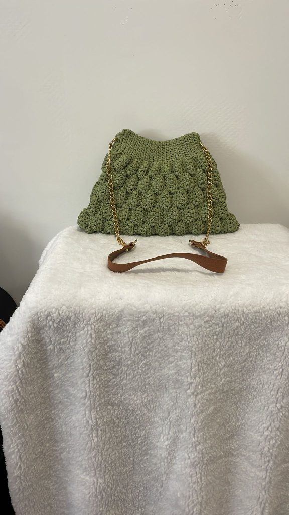 Sac à main trapèze en crochet couleur vert sauge avec une texture en relief point bulle ou noisette. Le sac est doté d'une anse en chaîne dorée et d'un renfort épaule marron. Création artisanale tendance Vanda sur fond blanc.