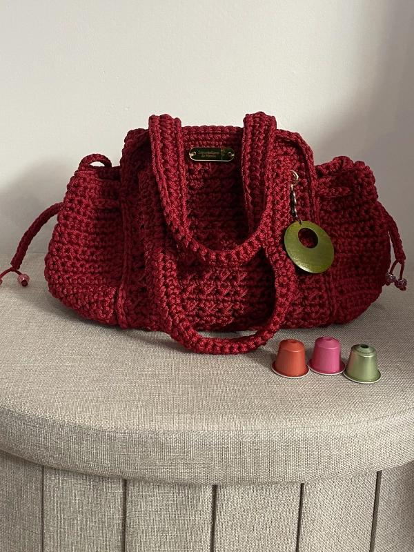 Sac à main au crochet rouge bordeaux fait main par Vanda, avec anses tressées et pompon en bois, posé sur un pouf en tissu